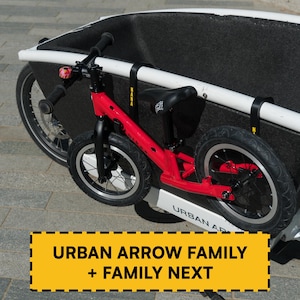 Puede incluir: Una bicicleta de equilibrio roja con ruedas negras y un timbre rosa en el manillar, montada en el lateral de una gran bicicleta de carga blanca y negra. La bicicleta de carga lleva el texto URBAN ARROW FAMILY + FAMILY NEXT en el cuadro.