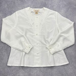 May include: A white long-sleeved blouse featuring a button-down front with five matching white buttons. The neckline is adorned with intricate white embroidery and cutwork detailing. A label reading MIKADO is visible at the inner collar.