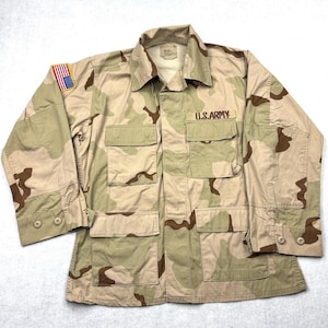 May include: A long-sleeve military-style jacket in a tan, brown, and beige desert camouflage pattern. The jacket features a collar, buttoned cuffs, four front pockets with flaps, and the text U.S. ARMY embroidered above the right pocket.