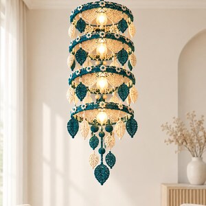 May include: A tiered macrame chandelier featuring four circular rings in deep teal, adorned with small white crocheted flowers and alternating teal and cream-colored leaf-shaped pendants. The fixture holds four warm-toned light bulbs.