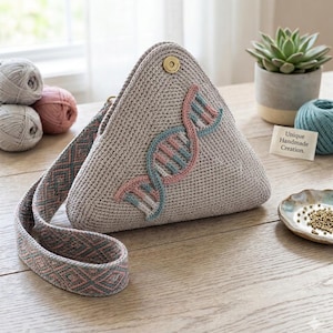 May include: A triangular crochet bag in light beige featuring a DNA double helix design embroidered in pink and blue. The bag has a matching woven strap with a geometric diamond pattern and a gold-toned magnetic snap closure at the top point.