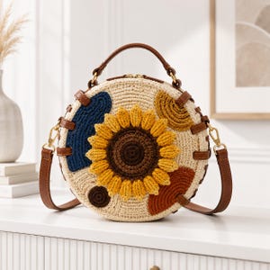 May include: A round, woven straw handbag featuring a large, textured, crocheted sunflower in the center. The bag is accented with brown leather straps, gold-tone hardware, and abstract crocheted shapes in navy blue, mustard yellow, and rust orange.