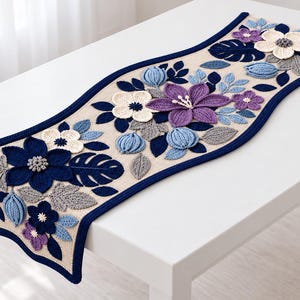 Könnte beinhalten: Ein beigefarbener Tischläufer mit dunkelblauem Rand und einem komplexen Häkel-Blumenmuster. Das Design zeigt dreidimensionale Blüten und Blätter in Marineblau, Hellblau, Violett, Creme und Grau, die über den Stoff verteilt angeordnet sind.