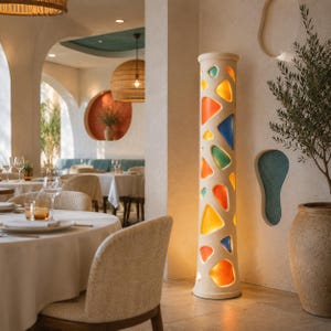 May include: A tall, cylindrical floor lamp with a textured off-white exterior featuring irregular, cut-out openings. These openings are filled with translucent, multi-coloured glass inserts in shades of orange, yellow, blue, and green.