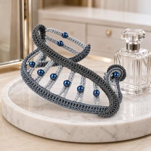May include: A hair claw clip featuring a dark gray crocheted frame with light gray crocheted accents and small, round, glossy dark blue beads embedded along the structure. The clip has a curved, open design with a sturdy, functional mechanism.