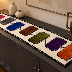 Pode incluir: Um caminho de mesa retangular com base na cor creme e borda costurada em preto. Apresenta uma série de seis retalhos retangulares de textura aveludada em cores vibrantes: magenta, cinza escuro, azul, marrom, verde oliva, roxo e laranja.