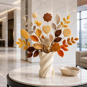 Puede incluir: Un arreglo decorativo de hojas otoñales y flores con forma de galleta tejidas a ganchillo en tonos marrón, amarillo y crema. Los tallos se sostienen en un jarrón blanco de columna retorcida, creando una muestra texturizada de arte textil.