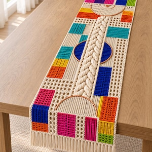 Pode incluir: Um caminho de mesa retangular em macramé com um cordão trançado central ladeado por dois aros semicirculares cor de cobre. O design incorpora uma grelha de cordão creme intercalada com blocos vibrantes em rosa, azul, laranja, amarelo e verde.