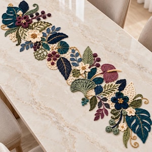 Puede incluir: Un camino de mesa decorativo largo con intrincados trabajos de ganchillo en un tema botánico. El diseño incluye varias hojas, flores y bayas en tonos azul profundo, verde bosque, crema y ciruela oscuro, dispuestos en un patrón continuo.