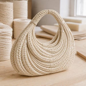 May include: A crescent-shaped handbag crafted from off-white macrame cord. The bag features a structured, rounded handle and a series of concentric, curved rows of braided rope that form the body, secured with small, matching cord wrappings.