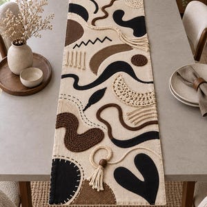 May include: A rectangular table runner featuring a cream-colored fabric base decorated with various abstract shapes in black, dark brown, and tan. The design includes textured yarn elements, crochet circles, fringe tassels, and stitched outlines.