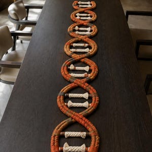 May include: A decorative table runner shaped like a DNA double helix, constructed from braided orange and brown fabric strands with white rope cross-links, laid out in a repeating spiral pattern across the length of a dark wooden table surface.