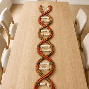 May include: A macrame wall hanging shaped like a DNA double helix structure. The piece features two long, twisted strands in shades of rust, orange, and brown, connected by horizontal rungs made of thick, off-white braided cotton cord.