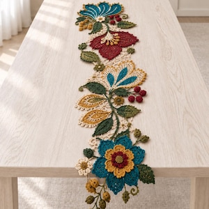 Puede incluir: Un camino de mesa de ganchillo largo y estrecho con un vibrante patrón floral. El diseño incluye flores intrincadas y multicolores en tonos azul verdoso, rojo intenso, amarillo mostaza y verde oliva, con pequeñas bayas y hojas.