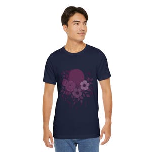 May include: Navy blue t-shirt featuring a floral design in shades of purple. The graphic includes roses, other flowers, and leaves, with a circular shape in the background. The shirt is a classic crew neck style.