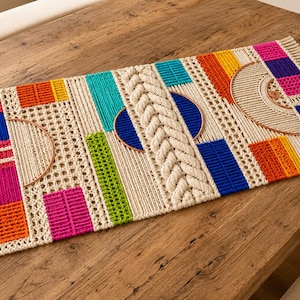 May include: A rectangular macrame table runner featuring a cream-colored base with fringe on the ends. It incorporates vibrant geometric blocks in pink, orange, blue, turquoise, and lime green, along with a central braided rope and two wooden hoops.