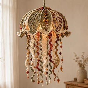 Puede incluir: Una lámpara colgante de ganchillo con una pantalla en forma de cúpula, decorada con patrones de hojas detallados en tonos verde oliva, crema y óxido. Numerosos zarcillos largos y en espiral cuelgan hacia abajo, adornados con pequeñas cuentas.