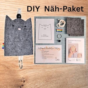 Könnte beinhalten: Ein DIY-Nähset für einen katzenförmigen Schlüsselanhänger. Das Set enthält grauen Filz, eine Vorlage, Anleitungen und weiteres Zubehör. Der Text "DIY Näh-Paket" befindet sich oben im Bild.
