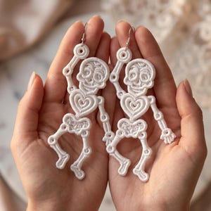 Può includere: Orecchini bianchi a forma di scheletro con intricati dettagli in pizzo. Gli orecchini presentano un teschio, una gabbia toracica a forma di cuore e arti articolati. Gli orecchini sono tenuti in mani aperte, mostrando il design delicato.