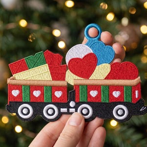 Könnte beinhalten: Ein gestickter Aufnäher in Form eines Eisenbahnwagens mit Geschenken und Herzen. Der Zug ist rot und grün mit weißen Herzakzenten und schwarzen Rädern. Er trägt rot und grün verpackte Geschenke, ein großes rotes Herz und eine blaue Schlaufe.