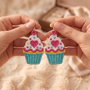 Könnte beinhalten: Ein Paar bestickte Cupcake-Ohrringe mit einer türkisfarbenen Basis, einem weißen Zuckerguss mit einem rosa Herz und bunten Punkten. Die Ohrringe sind an silberfarbenen Metallhaken befestigt, die das detaillierte Stickmuster hervorheben.