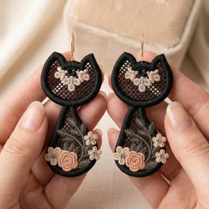 Könnte beinhalten: Ein Paar schwarze Katzenohrringe mit floraler Stickerei. Die Ohrringe haben eine schwarze Kontur mit einem netzartigen Design und sind mit beigen und rosa Blumen verziert. Sie werden von goldfarbenen Haken gehalten.