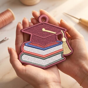 May include: A maroon embroidered patch featuring a graduation cap resting on a stack of three books. The books have white pages with blue, red, and grey covers. A gold tassel hangs from the cap, and a circular loop is at the top for hanging.