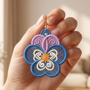 Könnte beinhalten: Nahaufnahme eines blütenförmigen Ohrrings, der in einer Hand gehalten wird. Der Ohrring hat ein blaues, lila, gelbes und weißes Stickdesign. Der Ohrring ist aus Stoff und hat einen silberfarbenen Haken.