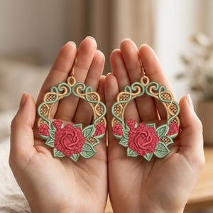 Könnte beinhalten: Gestickte Creolen mit einem Rosendesign. Die Ohrringe haben einen goldfarbenen Rahmen mit einem grünen und goldenen Filigranmuster. Am unteren Rand jedes Ohrrings ist eine rote Rose mit grünen Blättern gestickt.