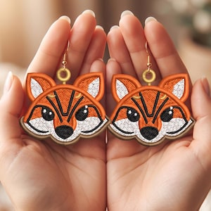 Könnte beinhalten: Ein Paar bestickte Ohrringe in Form von Fuchsköpfen mit Details aus orangefarbenem, weißem und schwarzem Garn. Jedes Fuchsgesicht hat große schwarze Augen mit weißen Glanzlichtern, eine schwarze Nase und spitze Ohren.