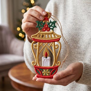 Könnte beinhalten: Ein dekorativer Hängeschmuck mit einem gestickten Laternendesign. Die Laterne ist goldfarben mit roten Akzenten und enthält eine weiße Kerze mit gelb-roter Flamme. Oben sind grüne Stechpalmenblätter und rote Beeren detailliert aufgestickt.