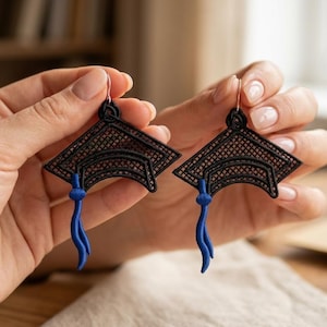 Könnte beinhalten: Ein Paar schwarze Abschlusskappen-Ohrringe mit blauen Quasten. Die Ohrringe werden von den Händen einer Person gehalten. Die Ohrringe sind ein einzigartiges Accessoire für Abschlussfeiern.