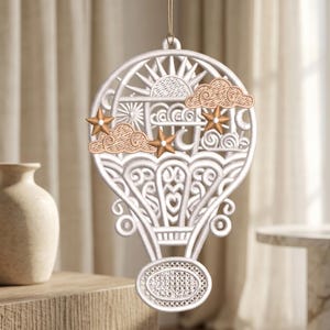 Könnte beinhalten: Ein weißes, besticktes Heißluftballon-Ornament mit komplizierten Ausschnittmustern. Das Design zeigt eine Sonne, eine Mondsichel und wirbelnde Wolken. Drei metallisch goldene Sterne sind auf der Vorderseite angebracht, und eine dünne Schnur dient zum Aufhängen.