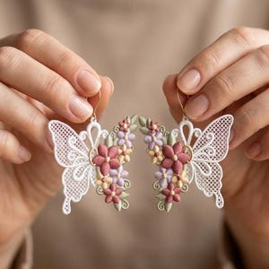 May include: Delicate white butterfly earrings with intricate lace-like wings and gold-colored hooks. The earrings are adorned with clusters of pink, purple, and yellow flowers. A beautiful accessory for a special occasion.