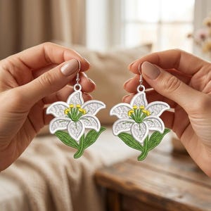 Könnte beinhalten: Ein Paar Ohrhänger mit gestickten weißen Lilienblüten, grünen Stielen und gelben Staubgefäßen. Das florale Design ist mit detaillierten weißen Fadenstichen auf den Blütenblättern und grünem Faden für die Blätter gefertigt und an silberfarbenen Ohrhaken befestigt.