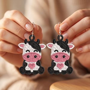Könnte beinhalten: Ein Paar Hängeohrringe mit gestickten Kuhmotiven. Jede Kuh ist weiß mit schwarzen Flecken, einer rosa Schnauze und einem kleinen rosa Herz auf der Brust. Die Ohrringe sind an silberfarbenen Metallhaken zum Einhängen in die Ohren befestigt.