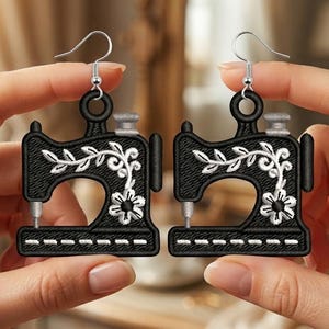 Könnte beinhalten: Ein Paar schwarze Ohrringe in Form von Vintage-Nähmaschinen mit weißer Blumenstickerei, einem silberfarbenen Knopf an der Oberseite und einer silberfarbenen Nadel, befestigt an silberfarbenen Metallhaken für durchstochene Ohrläppchen.