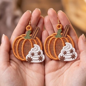 Könnte beinhalten: Ein Paar bestickte Kürbis-Ohrringe mit orangefarbenen Kürbisformen, grünen Stielen, einer braunen Zimtstange und einem weißen Klecks Schlagsahne, der mit kleinen braunen Sternanis-Akzenten verziert ist, befestigt an goldfarbenen Ohrhaken.