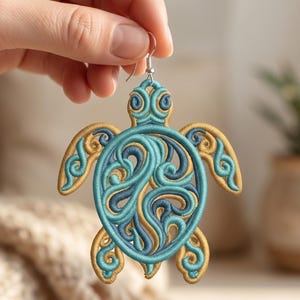 May include: Embroidered sea turtle earring with a teal and gold design. The turtle has a detailed, swirling pattern in the center and on its flippers. The earring is held up by a silver hook.