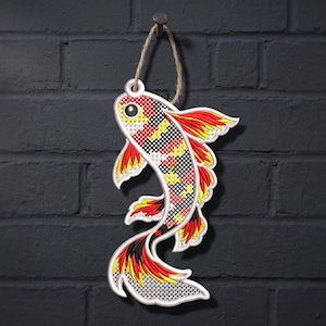 Könnte beinhalten: Ein hängendes Koi-Fisch-Ornament mit einem Kreuzstichmuster in Rot, Gelb, Schwarz und Weiß. Der Fisch hat einen weißen Rand, ein einzelnes schwarzes Auge und eine Juteschnur zum Aufhängen. Das Design zeigt ein strukturiertes Gittermuster.