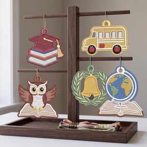 May include: A dark wood display stand holding five embroidered ornaments featuring school-themed designs: a graduation cap on books, a yellow school bus, an owl on a book, a bell in a wreath, and a globe on an open book, with embroidery floss below.