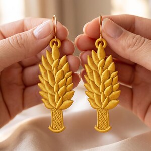 Könnte beinhalten: Ein Paar goldgelbe, bestickte Ohrhänger in Form von Weizenähren. Jeder Ohrring zeigt ein strukturiertes, gesticktes Design mit einem zentralen Stiel und geschichteten blattartigen Mustern, befestigt an einem einfachen goldfarbenen Haken.