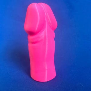 Peut inclure: Un objet phallique rose néon vif avec une finition mate et lisse. L'article présente une partie supérieure arrondie et une base cylindrique, posé à la verticale. La surface montre de fines lignes verticales typiques d'une impression 3D.