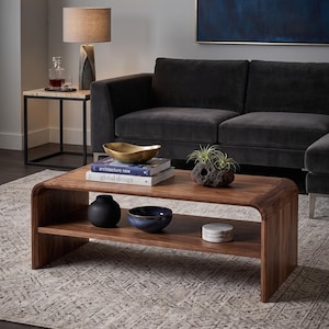 May include: A rectangular wooden coffee table with a curved waterfall edge design and a lower shelf. The top surface holds a gold bowl, two books titled Architecture Now and Global Design, and a small plant. The lower shelf holds a black vase, a blue bowl, and white coasters.