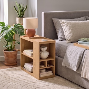 Puede incluir: Una mesita de noche de madera de roble claro con una superficie rectangular que incluye una base de lámpara integrada y una plataforma de carga circular. La unidad cuenta con dos estantes abiertos a la izquierda y compartimentos a la derecha con libros y un jarrón blanco.