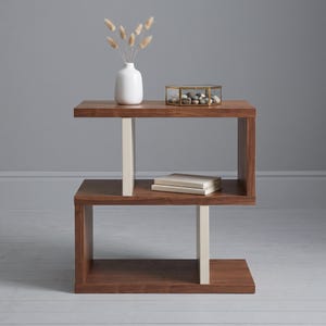 Puede incluir: Una mesa auxiliar de madera marrón con un marco en forma de S y dos soportes verticales de color crema. La superficie superior sostiene un jarrón blanco con tallos secos y una caja de cristal llena de piedras. Dos libros blancos están apilados en el estante central.