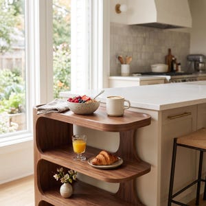 Puede incluir: Una estantería abierta de madera oscura de tres niveles con bordes redondeados. El estante superior contiene un cuenco moteado con fruta y granola con una cuchara y una taza color crema. El estante central contiene un vaso de zumo de naranja y un cruasán.