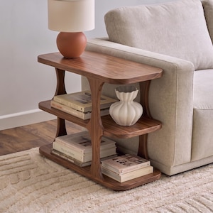 Puede incluir: Una mesa auxiliar de tres niveles hecha de madera de nogal con bordes redondeados y soportes verticales curvos. La mesa sostiene una lámpara color terracota, un jarrón blanco estriado y varios libros apilados en sus estantes.