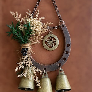 May include: A decorative wall hanging featuring a dark bronze-toned metal horseshoe suspended by a chain. Attached to the horseshoe are three brass-coloured bells, a circular pentagram charm, and a bundle of dried greenery tied with twine.