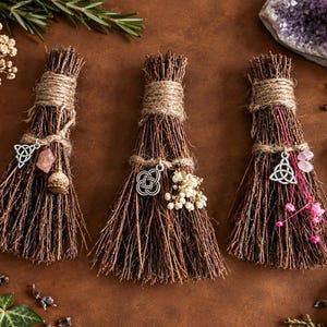May include: Three small decorative brooms made of bundled brown twigs, each tied with natural jute twine. Each broom features a silver-toned metal charm, including two triquetra symbols and one knot design, accented with small crystals and dried flowers.